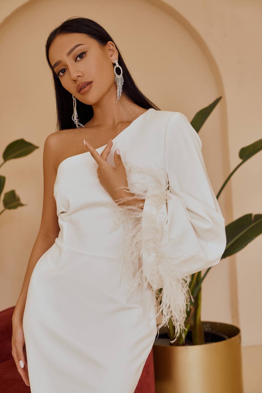 Woman in a white one-shoulder dress with fringe details, standing against a beige wall with plants.
