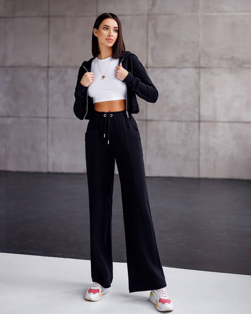 Woman wearing a black jacket, white crop top, and black wide-leg pants against a gray wall.