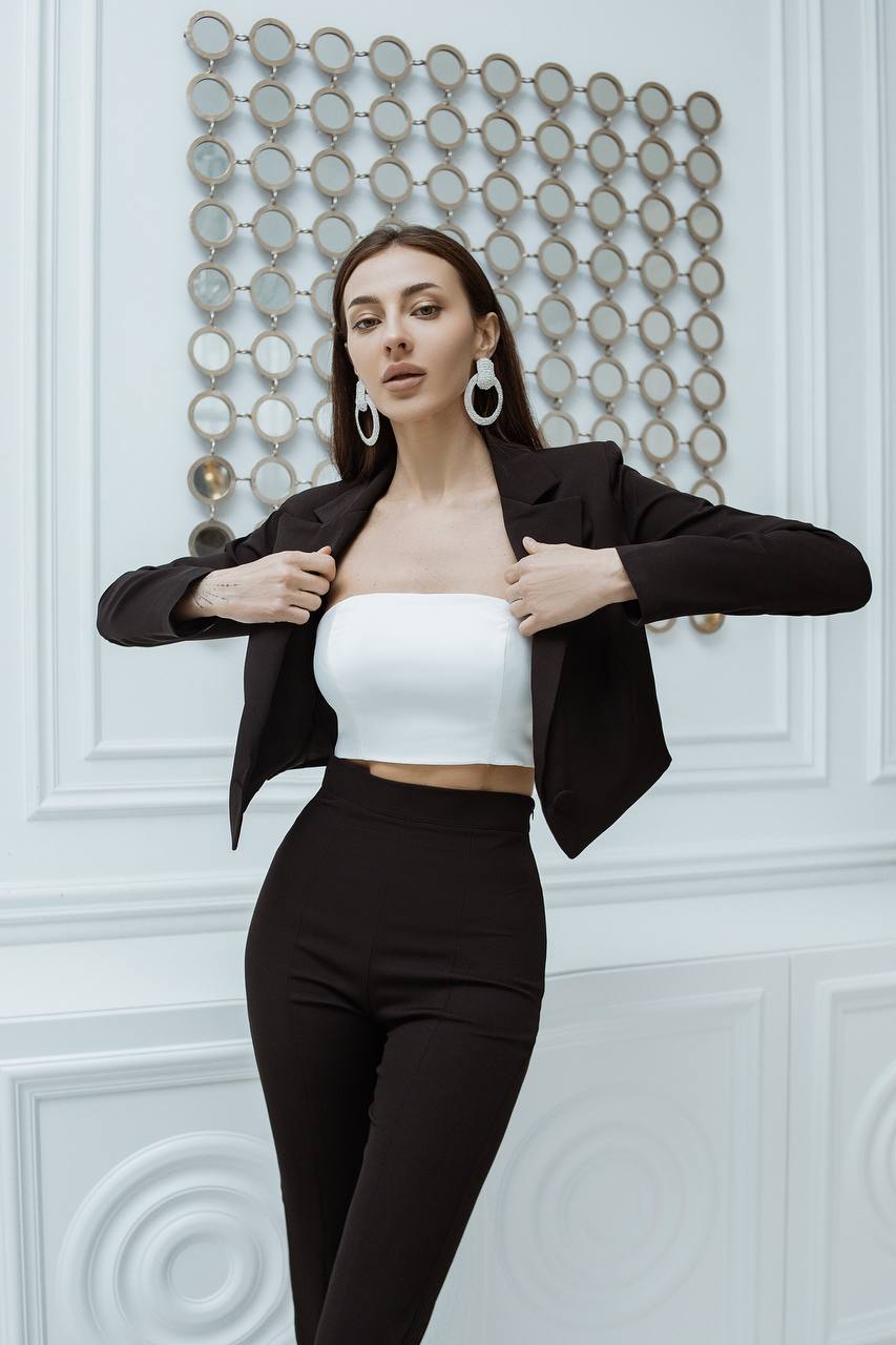 Woman wearing a black blazer over a white top and black pants against a decorative wall.