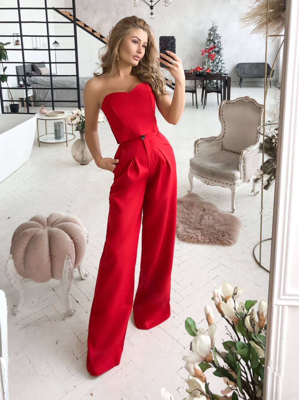 Woman in a red one-shoulder jumpsuit taking a mirror selfie in a stylish room.