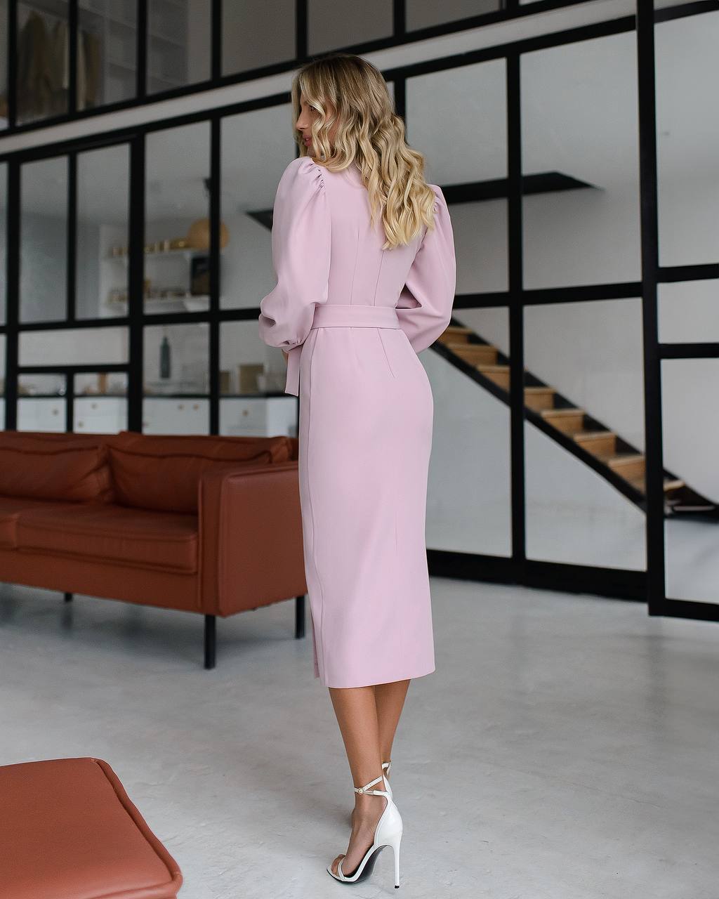 Woman in a pink dress standing in a modern interior setting with a brown sofa and glass walls.