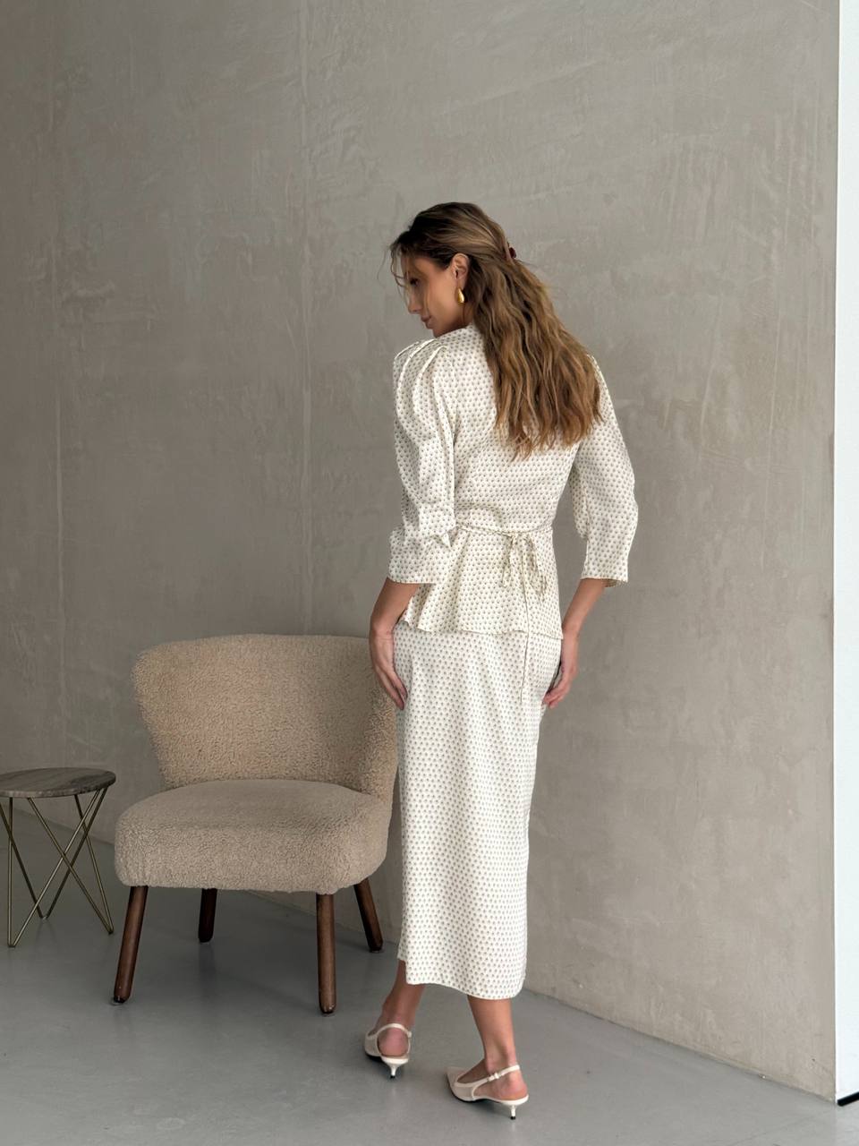 Woman in a white textured outfit standing in a minimalistic room with a chair and small table.