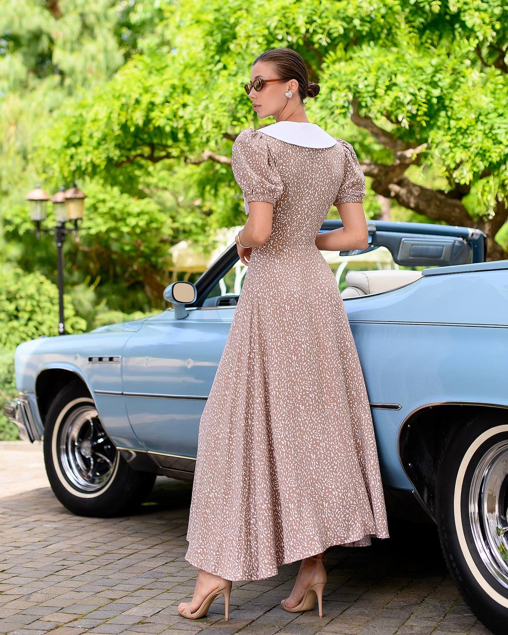 Woman in a vintage dress standing next to a classic car with greenery in the background