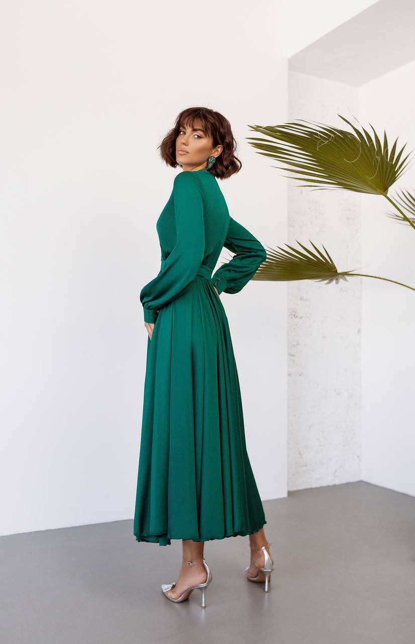 Woman wearing a green dress standing against a white wall with a plant.
