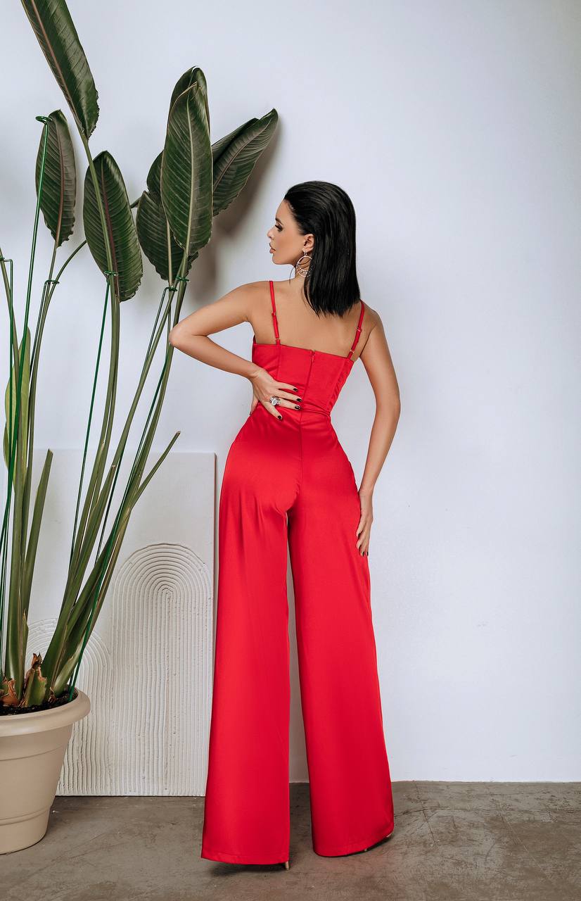 Woman in a red jumpsuit standing next to a potted plant against a white wall.