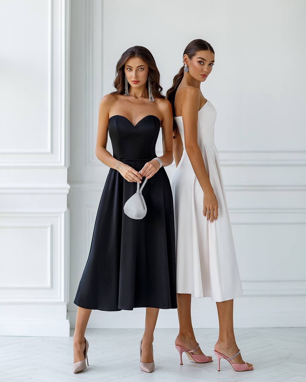 Two women wearing black and white dresses standing against a white wall.
