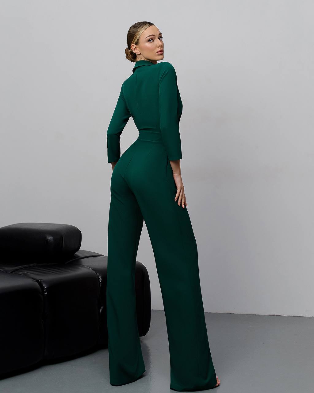Woman wearing a green jumpsuit standing in a minimalistic room with a black leather couch.