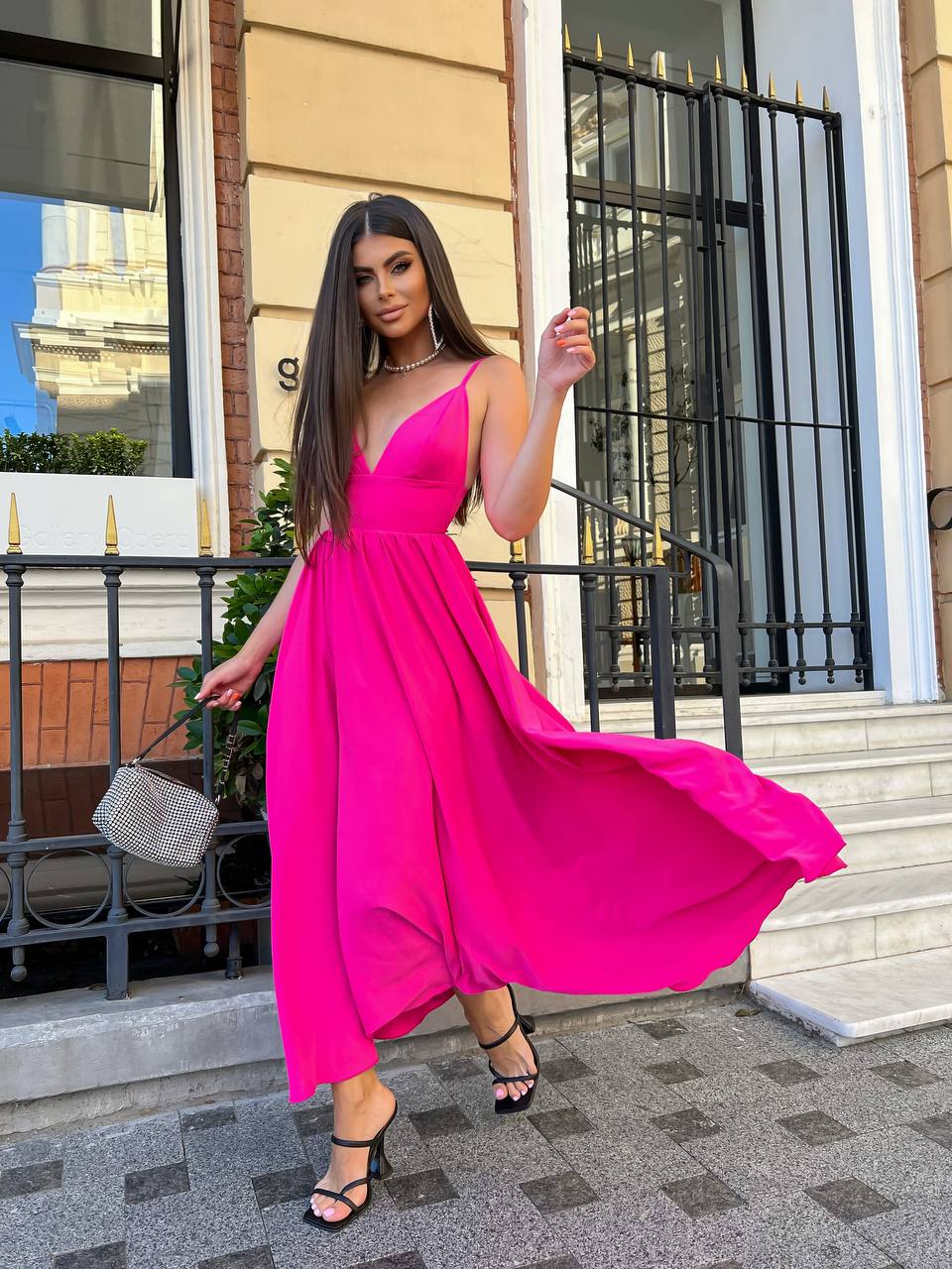 A woman wearing a bright hot pink sleeveless maxi dress with a V-neckline, standing on outdoor stone steps in front of a building with black metal railings.