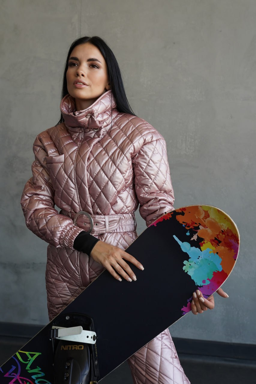 Woman in a pink quilted jacket holding a colorful snowboard against a gray background