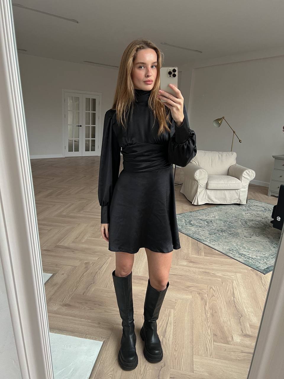 Woman taking a mirror selfie wearing a black outfit in a room with wooden flooring and a white armchair.