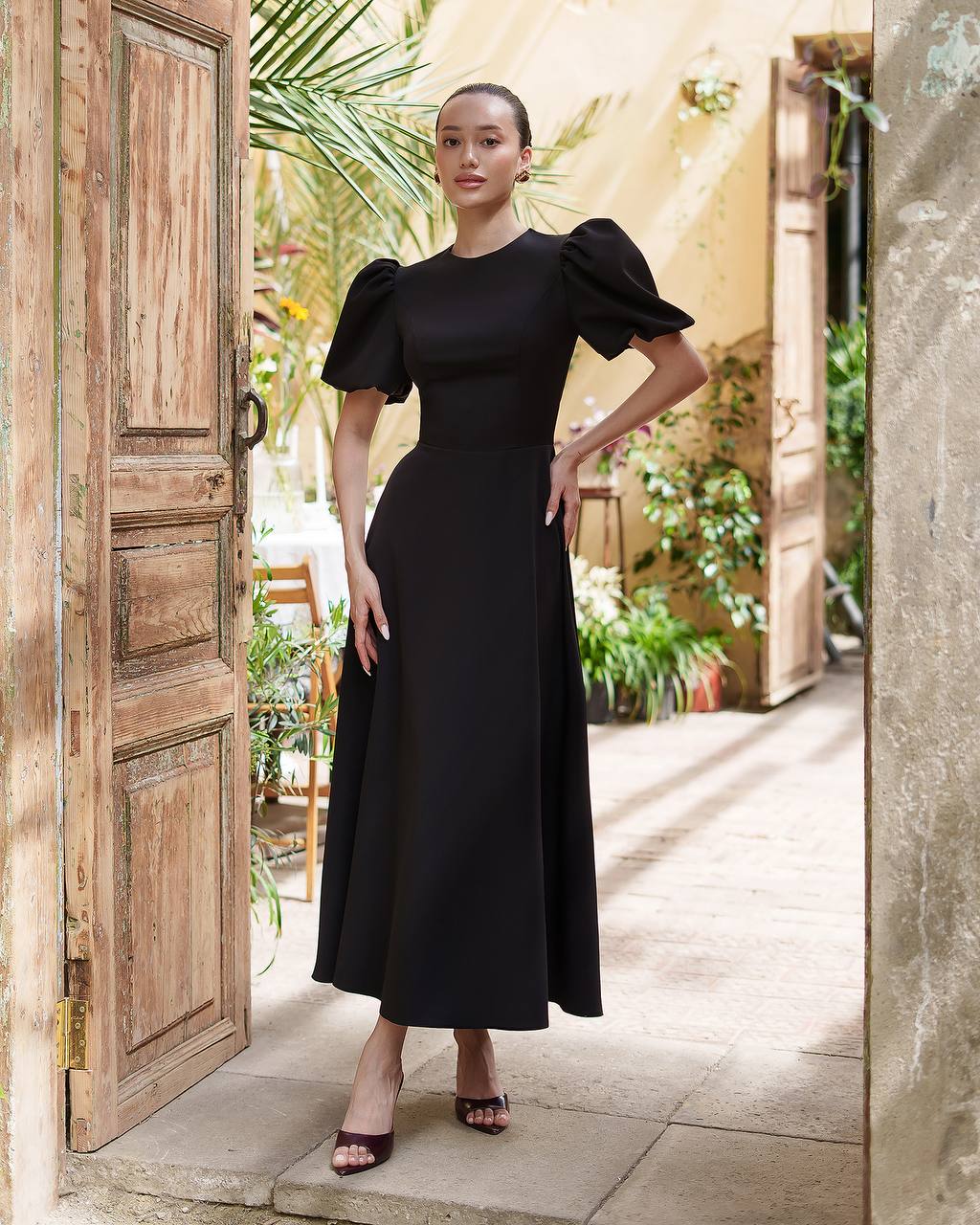 Woman in a black dress standing in an outdoor setting with plants and a wooden door.