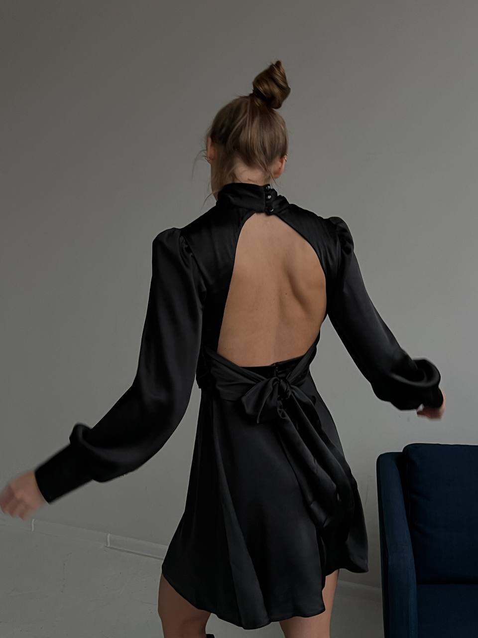 Woman wearing a black dress with long sleeves and an open back, standing against a plain background.