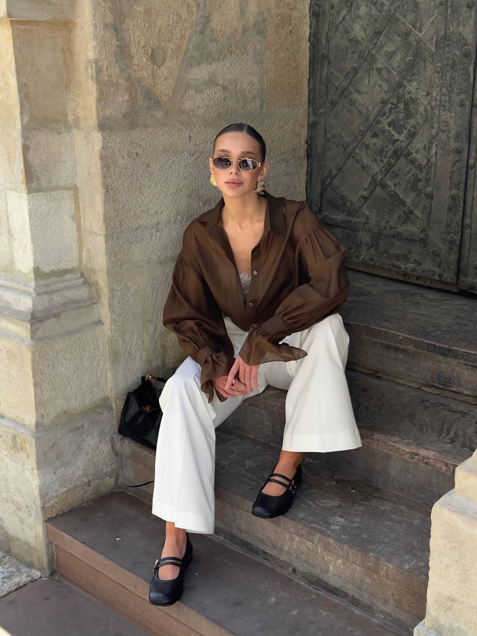 Woman sitting on stone steps wearing a brown blouse and white pants.