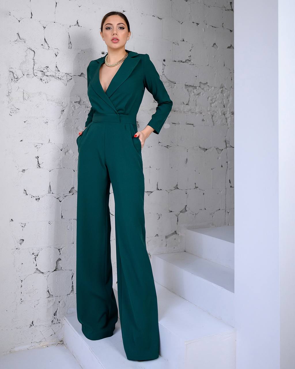 Woman wearing a green jumpsuit standing on white steps against a textured white wall.