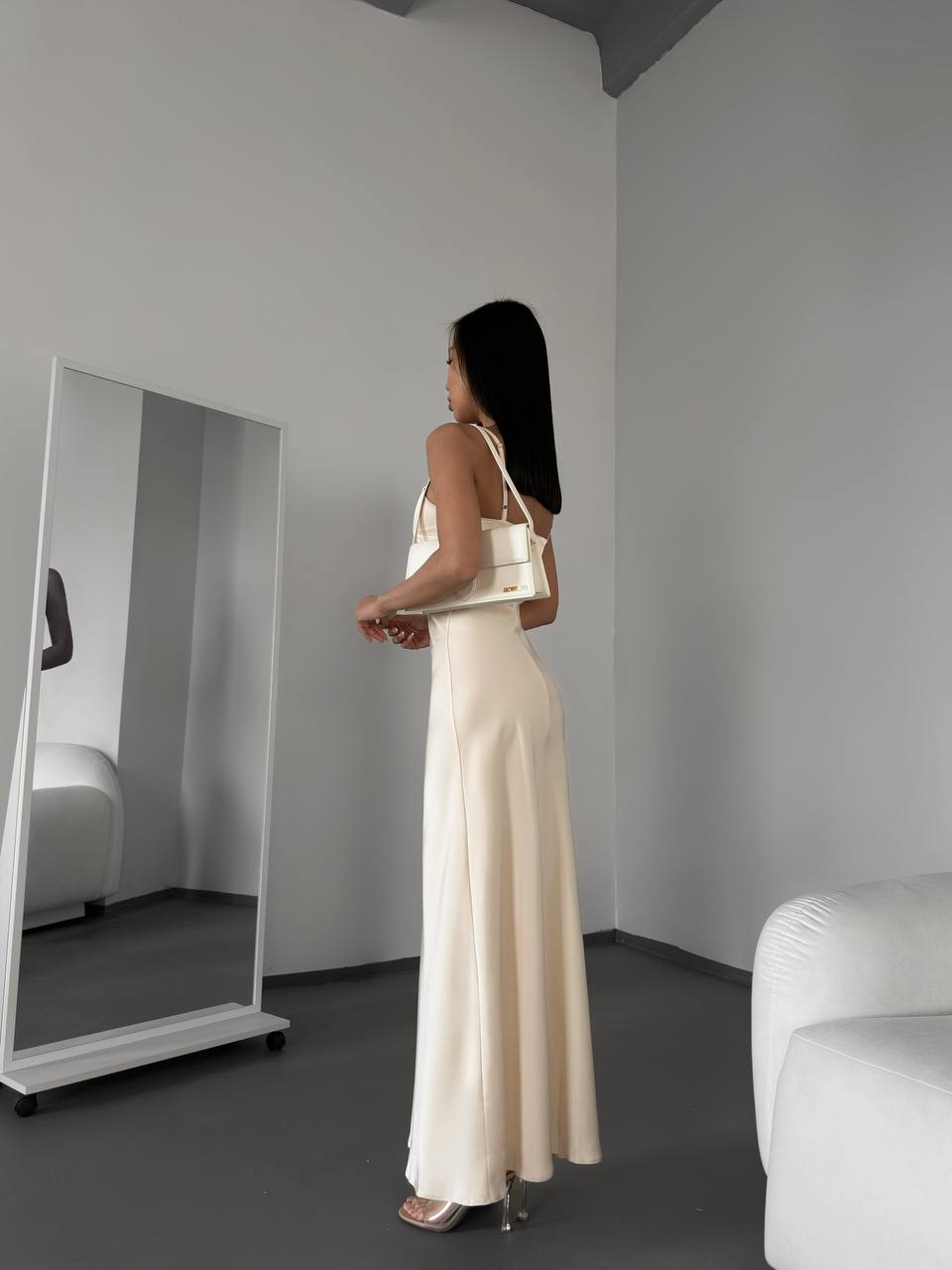 Woman in a long beige dress standing in a minimalistic room with a mirror.