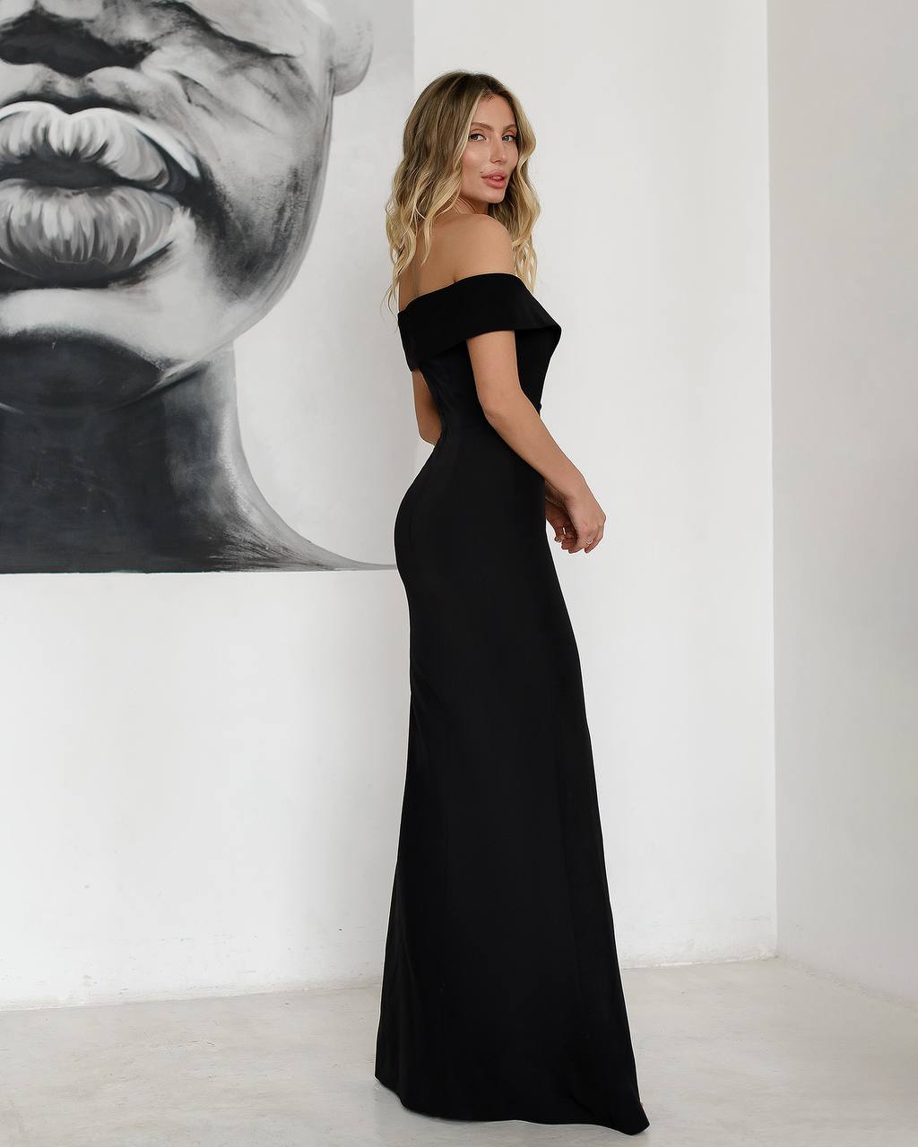 Woman wearing a black off-shoulder gown standing in a minimalistic room with a large black and white portrait on the wall.