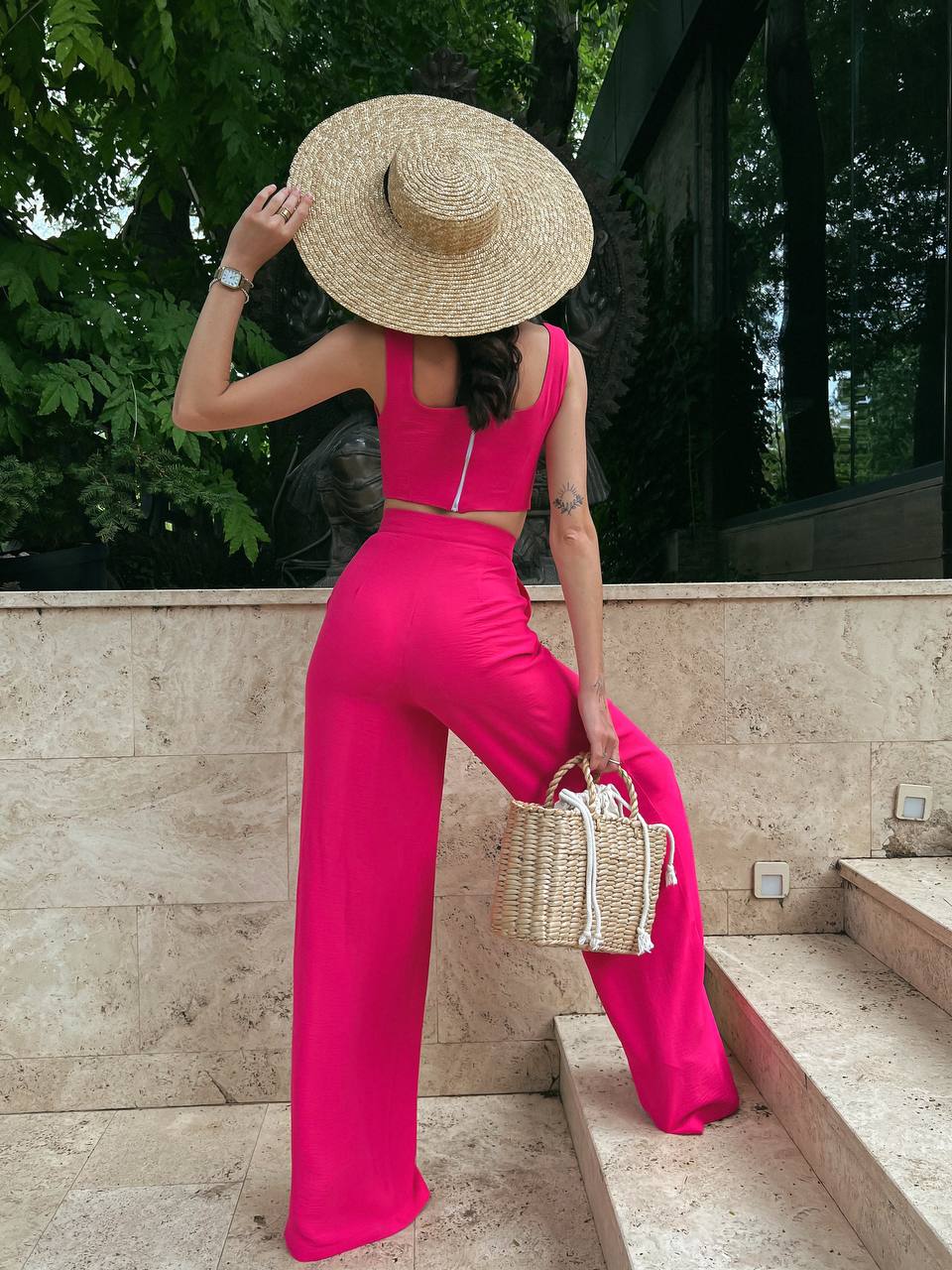 Woman in a bright pink outfit with a straw hat and bag, standing outdoors.