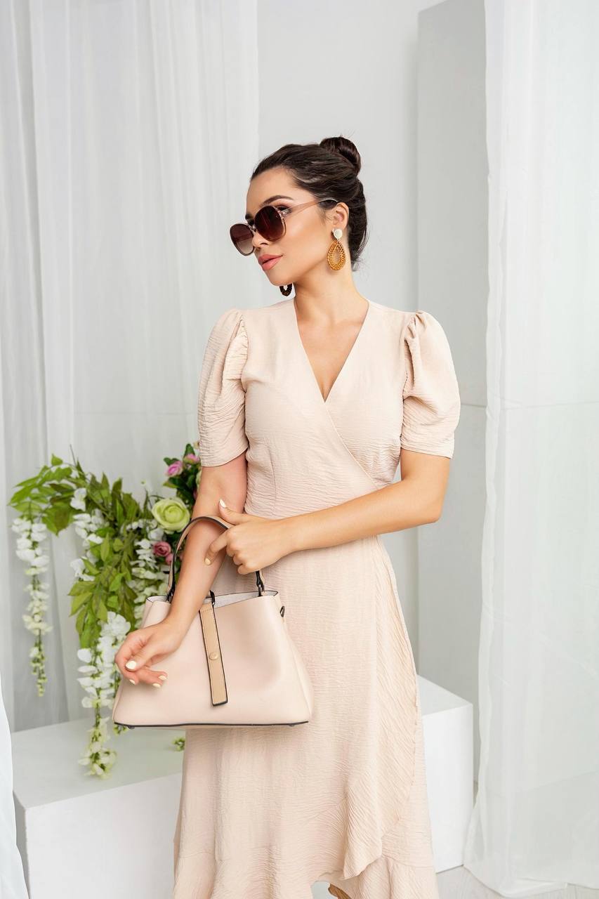 Woman in a beige dress holding a matching handbag against a white curtain background