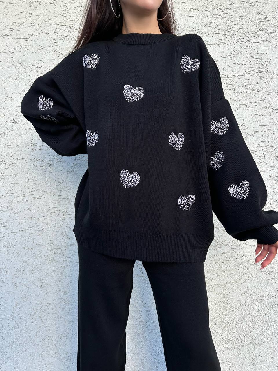 Black sweater with heart patterns worn by a person against a light gray textured wall.