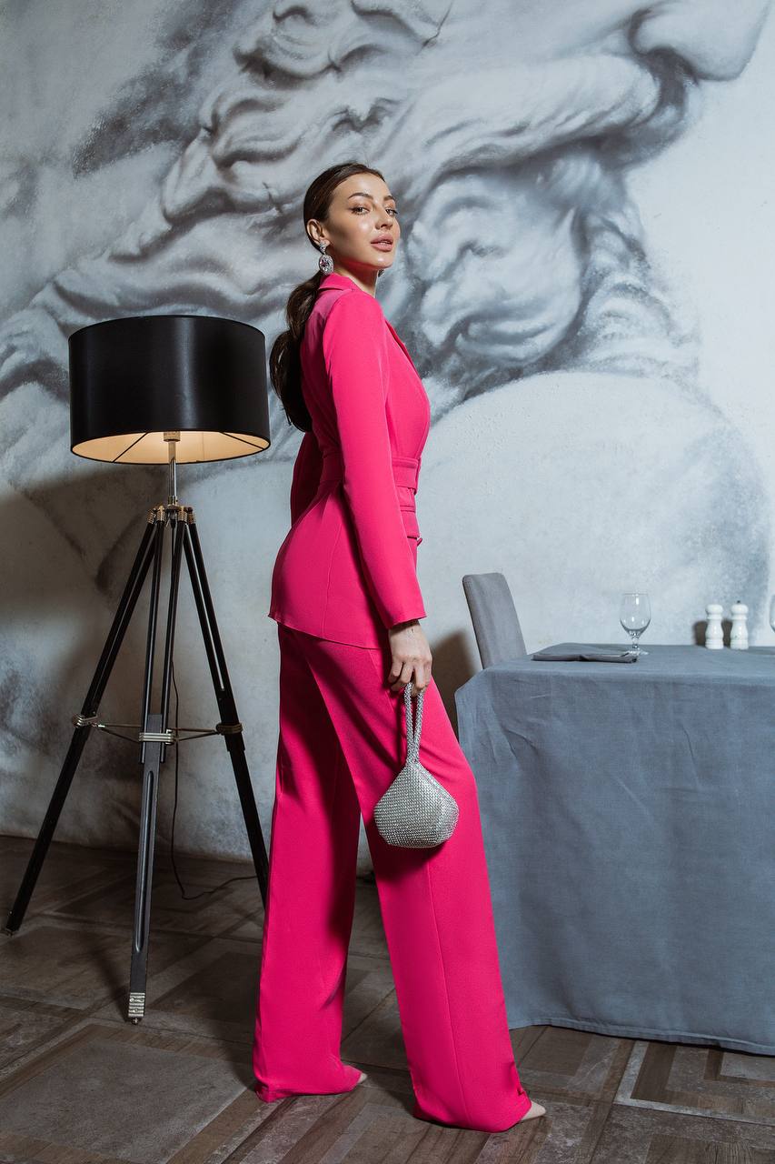 Woman in a bright pink outfit standing in a room with a lamp and table.