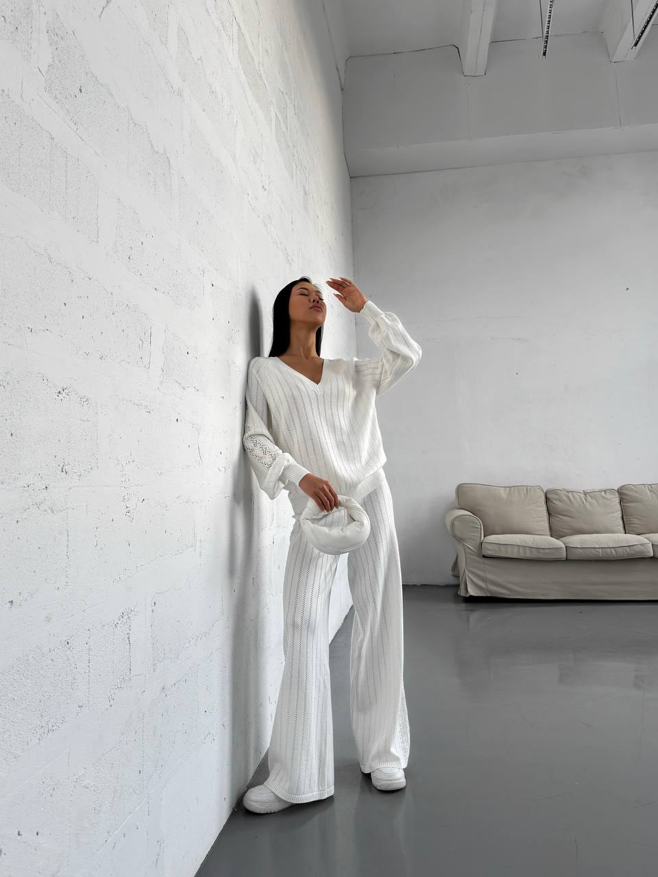 Woman in a white outfit standing against a white wall in a minimalistic room with a gray sofa.