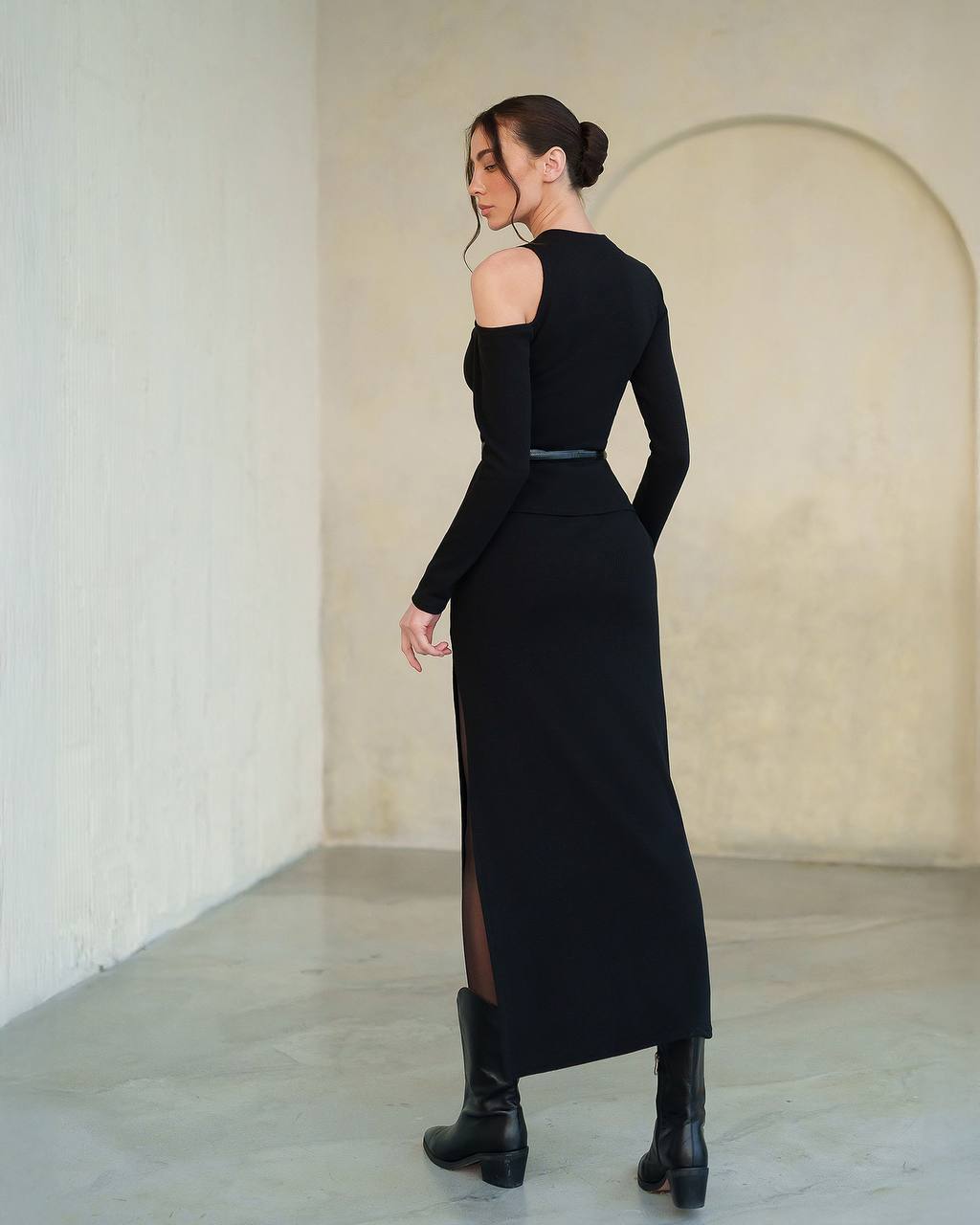 Woman wearing a black dress with long sleeves and a slit, standing in a minimalistic room.