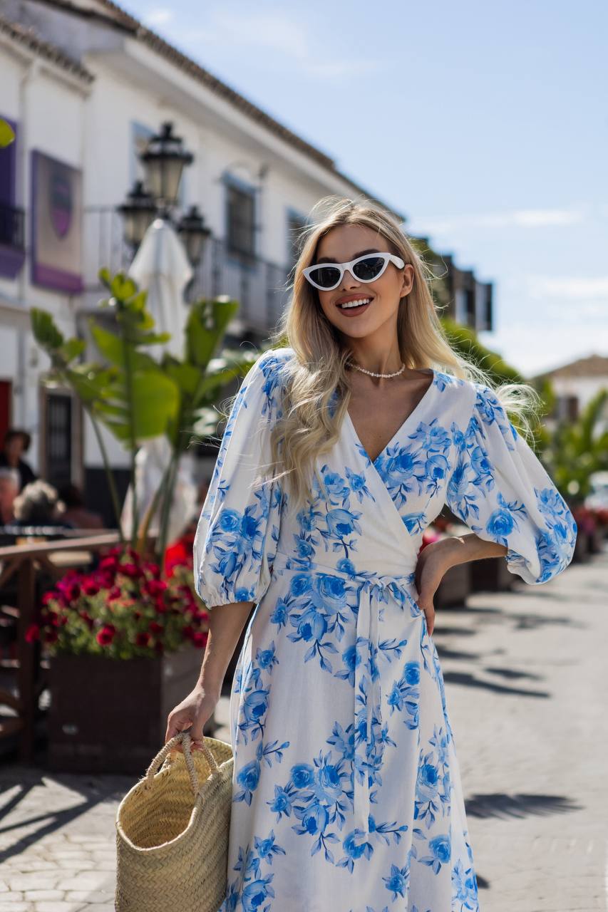 Woman in a blue floral dress walking on a street with buildings and plants in the background.