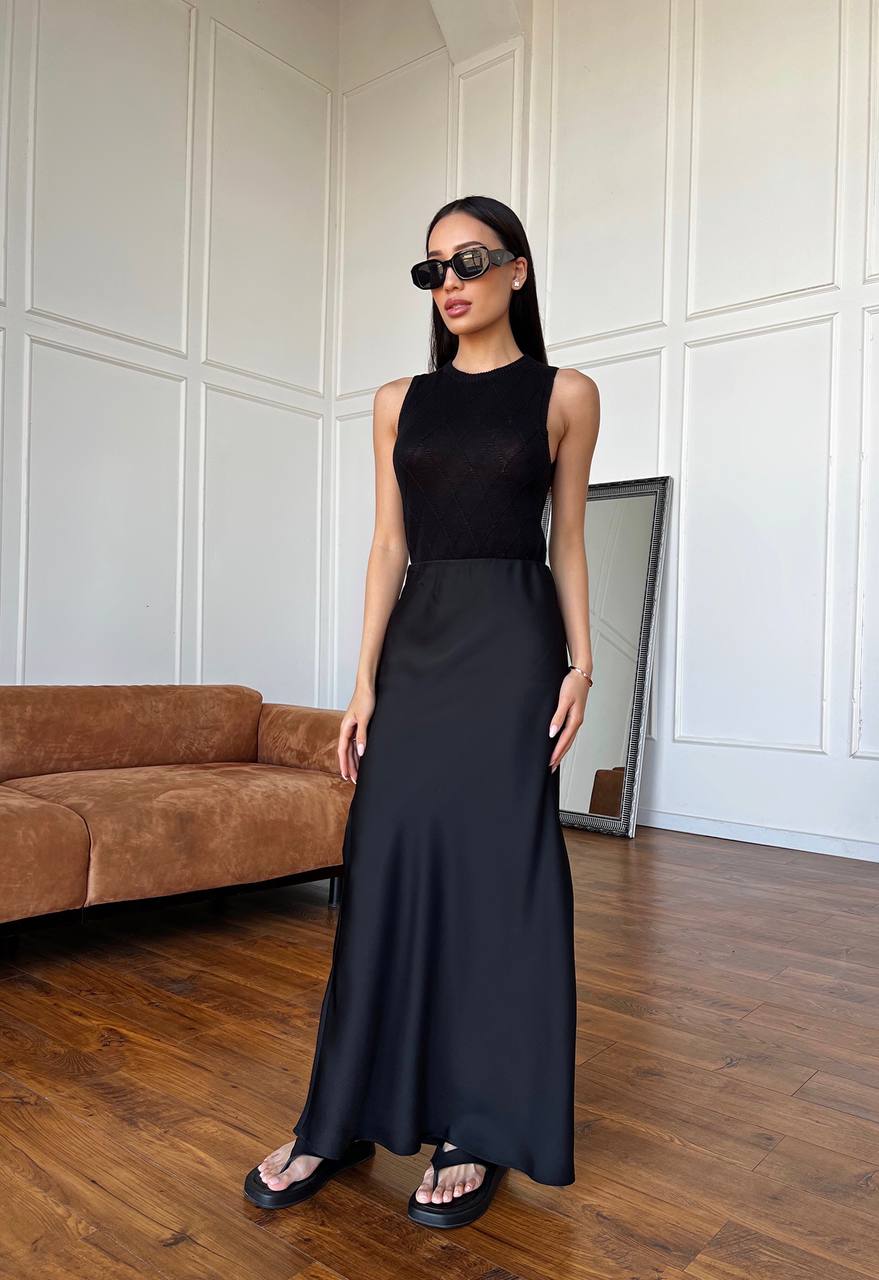 Woman in a black sleeveless top and long skirt standing in a room with a brown sofa and white paneled walls.