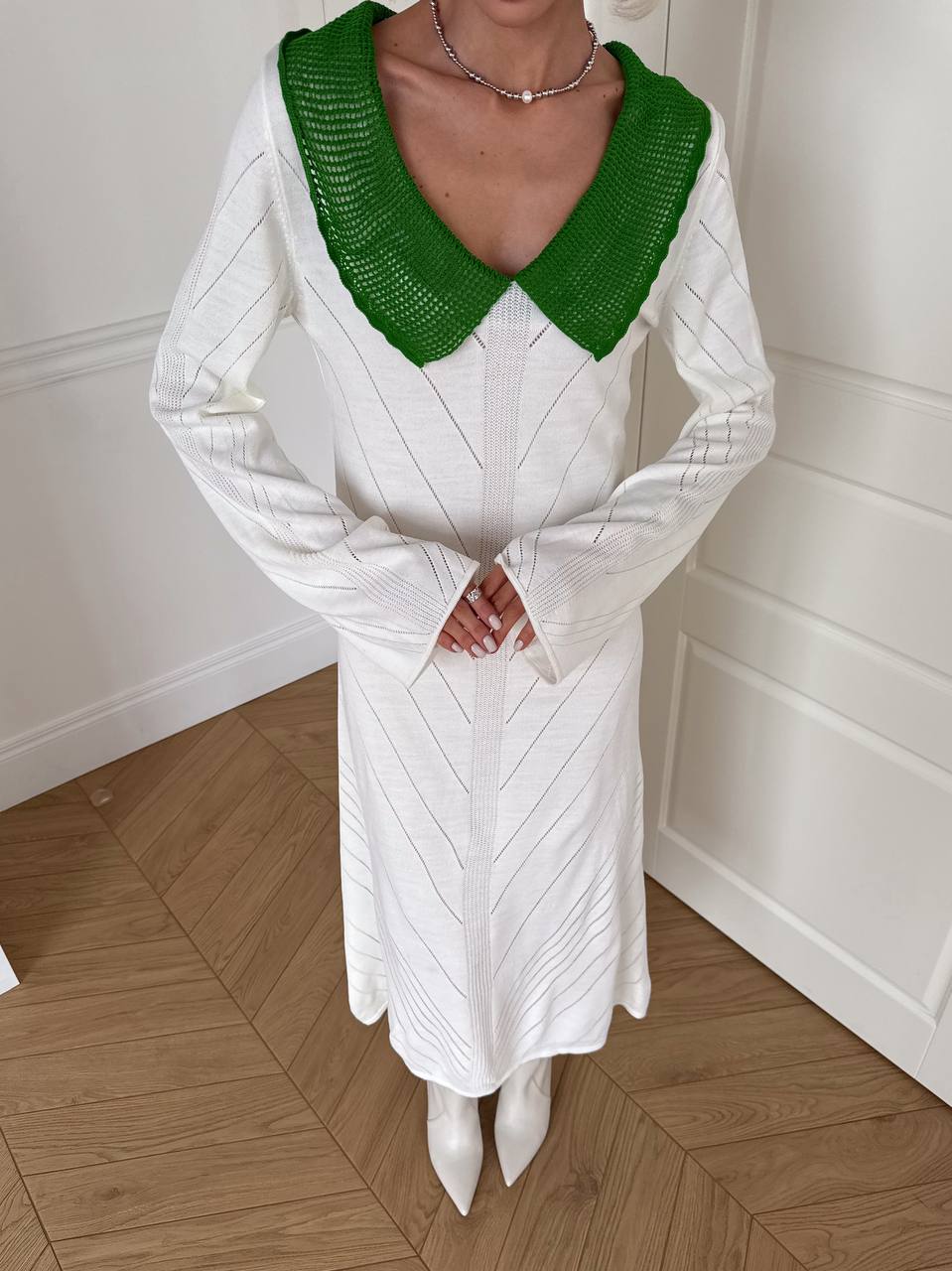 Person wearing a white dress with a green V-neck top against a white paneled wall.