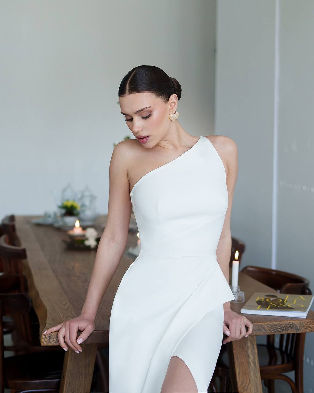 Woman in a white one-shoulder dress standing in a room with a table and chairs.