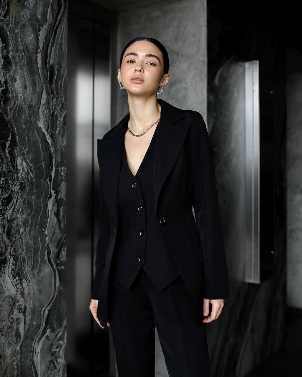 Person wearing a black suit standing against a dark, textured wall.