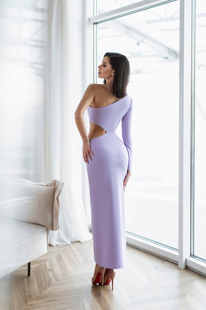 Woman in a lavender one-shoulder dress standing in a bright room with large windows.