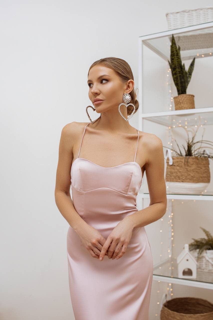 Woman wearing a pink satin dress with heart-shaped earrings in a room setting.