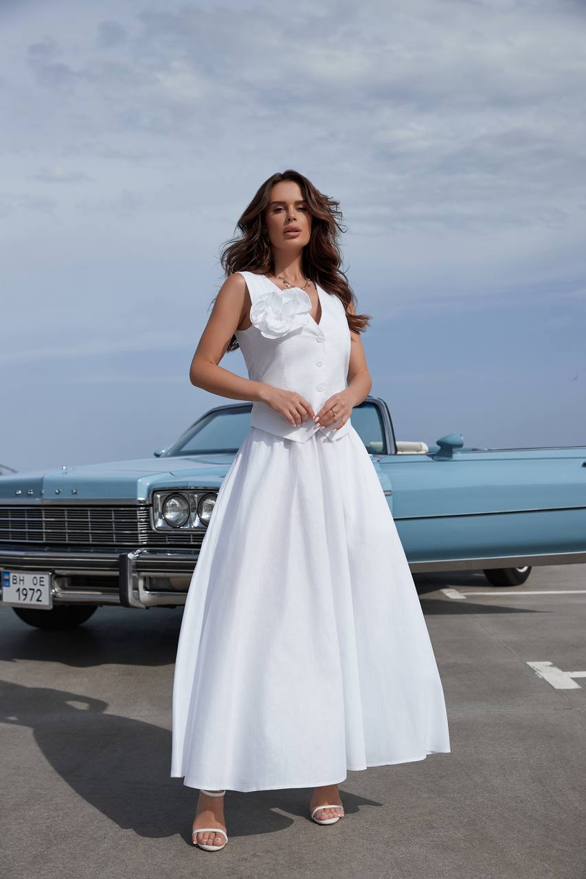 Woman in a white dress standing in front of a vintage blue car on a road.
