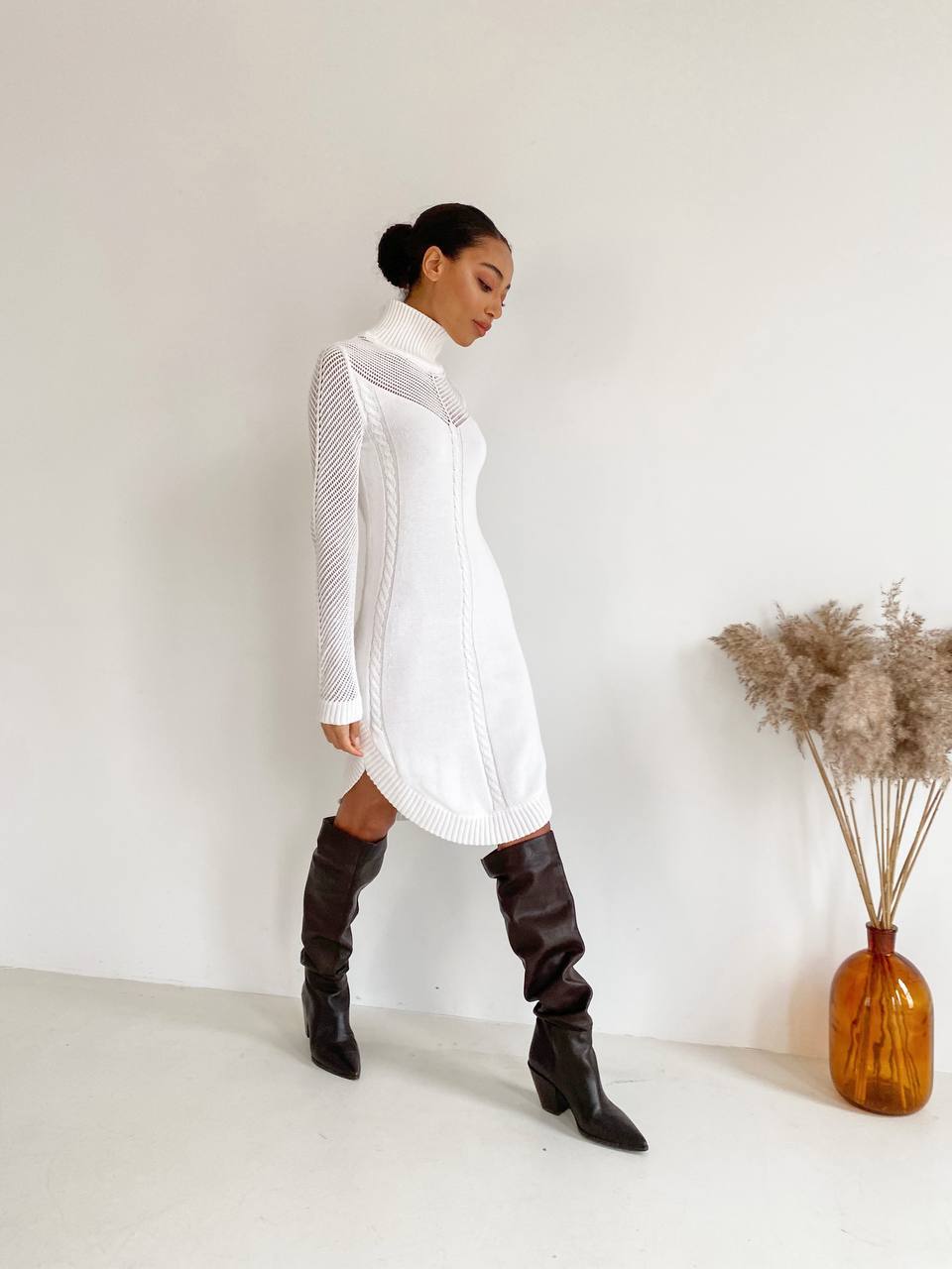 Woman wearing a white dress and knee-high boots in a minimalistic setting with a vase of dried plants.