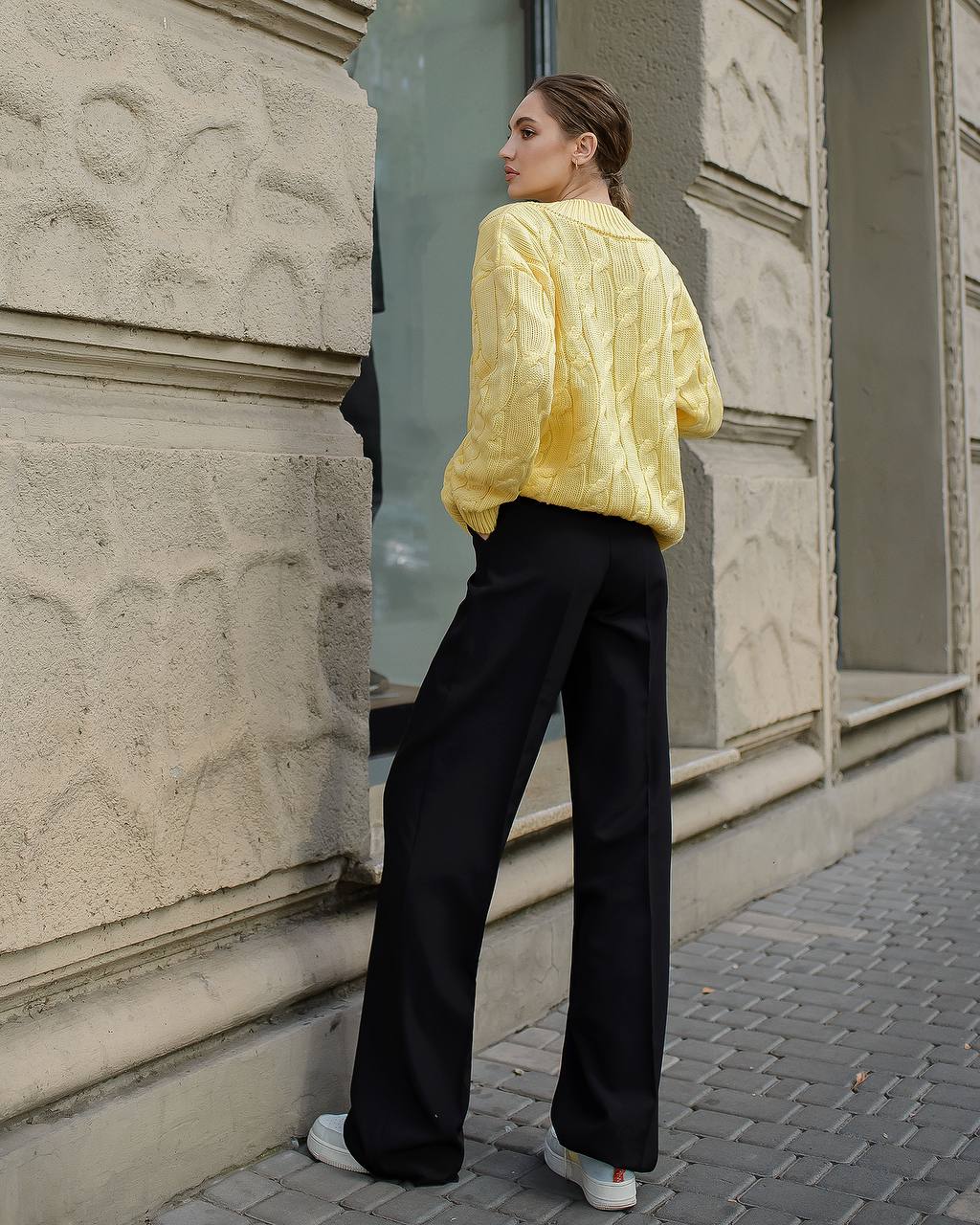 Woman wearing a yellow jacket and black pants standing against a stone building.