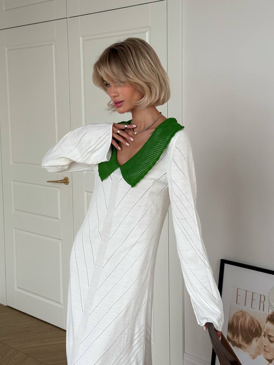 Woman wearing a white dress with a green neckline against a white wall.
