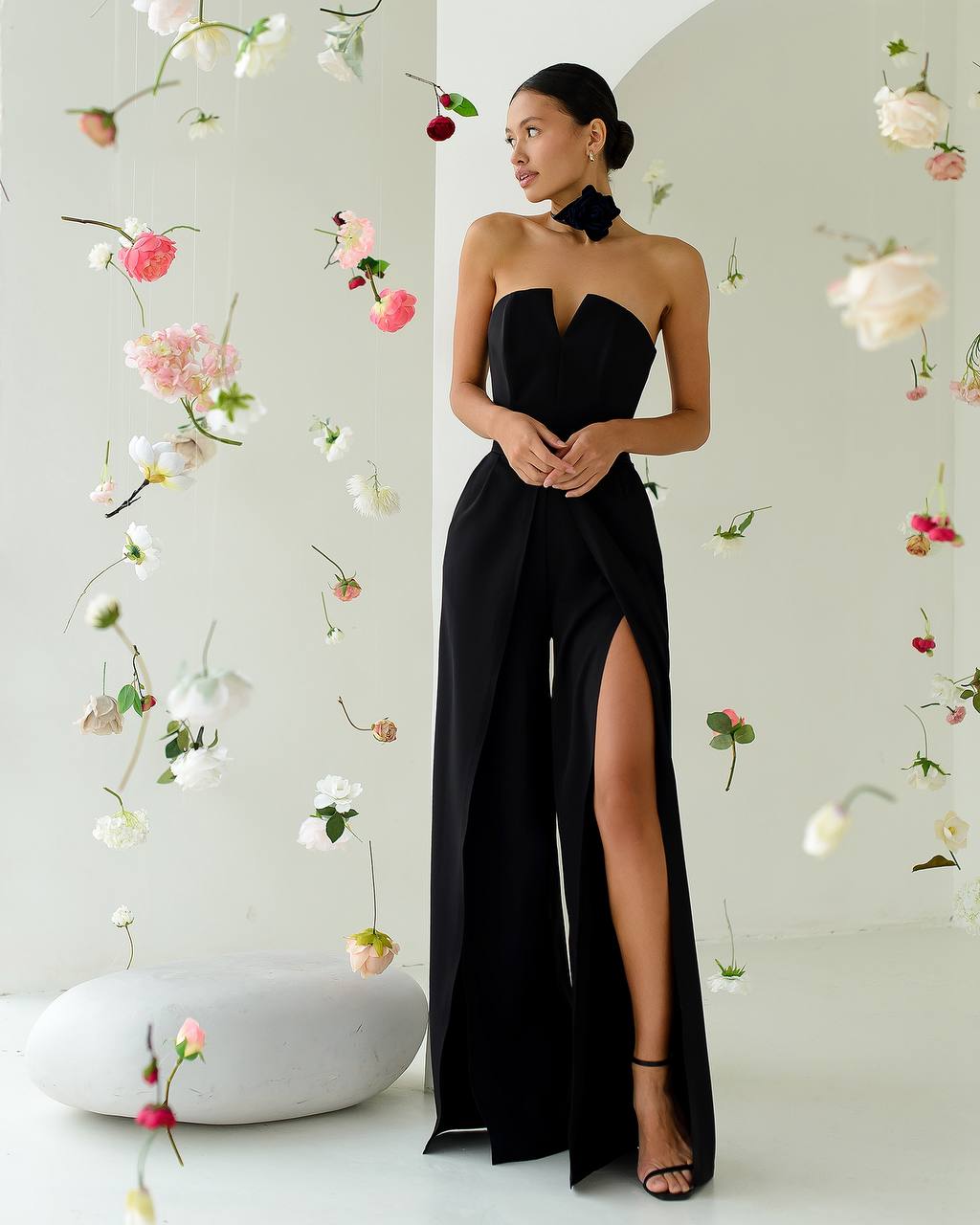 Woman in a black strapless gown with a high slit standing against a floral decorated wall.