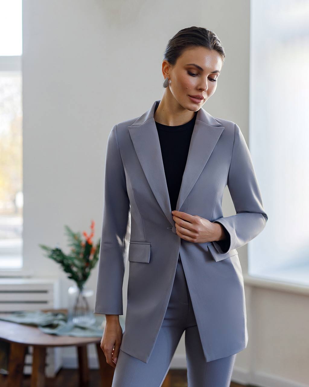 Woman wearing a light gray suit in a bright room with large windows.