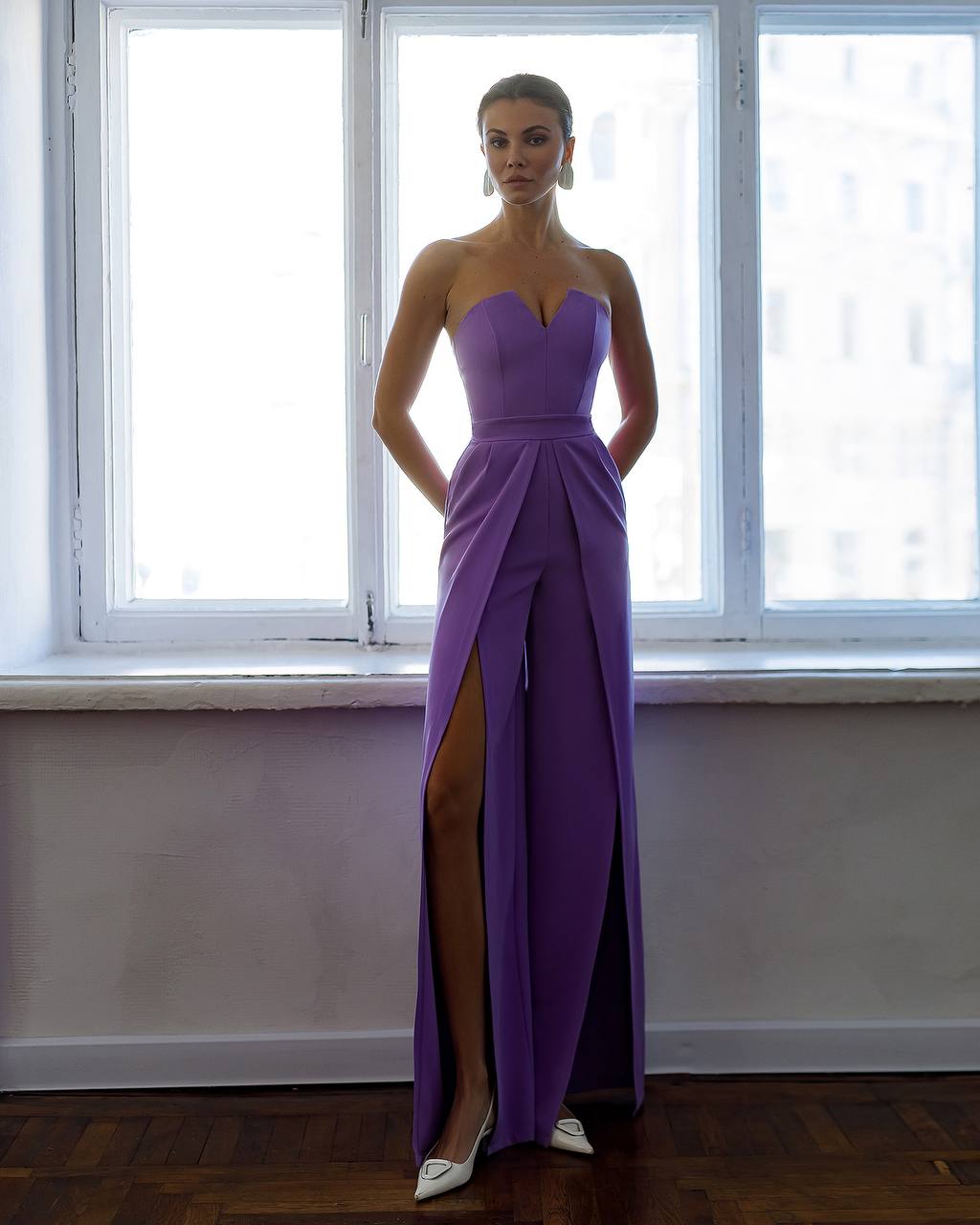 Woman wearing a strapless purple gown with a slit standing in front of a window.