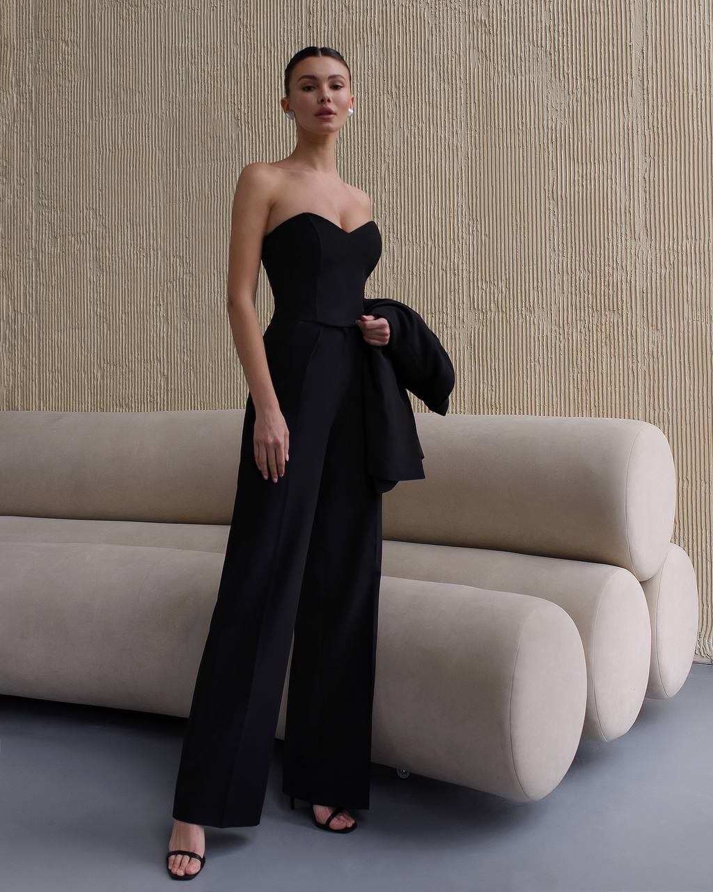 Woman in a black strapless outfit standing in front of a beige sofa against a textured wall.