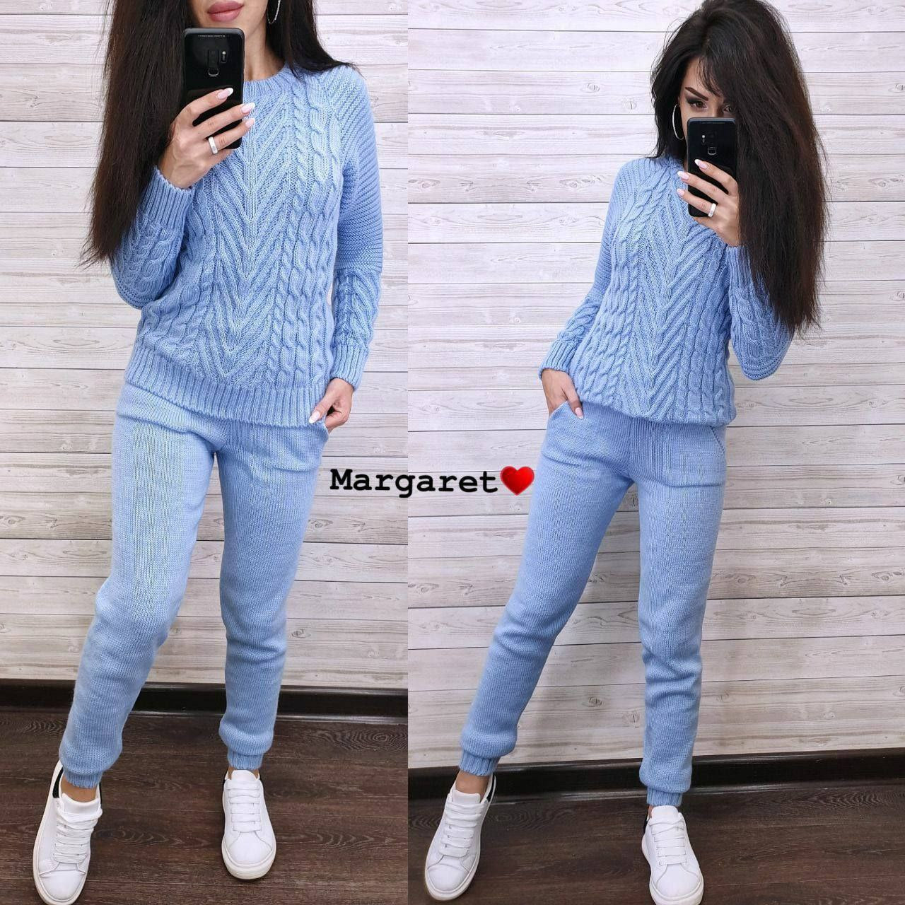 Woman wearing a blue cable knit sweater and pants set, taking a mirror selfie against a wooden panel background.