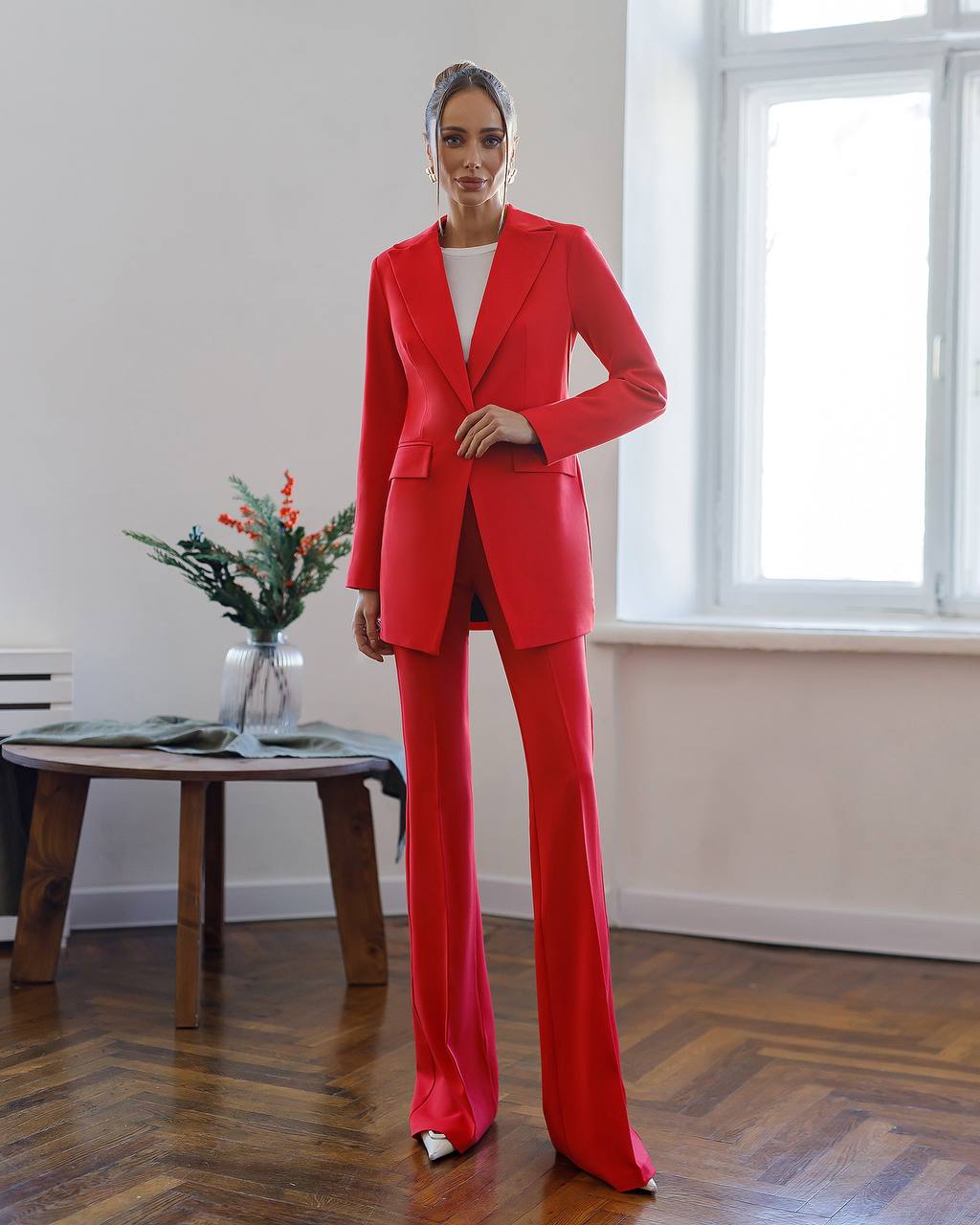 Person wearing a red suit standing in a room with a wooden floor and a vase of flowers on a table.