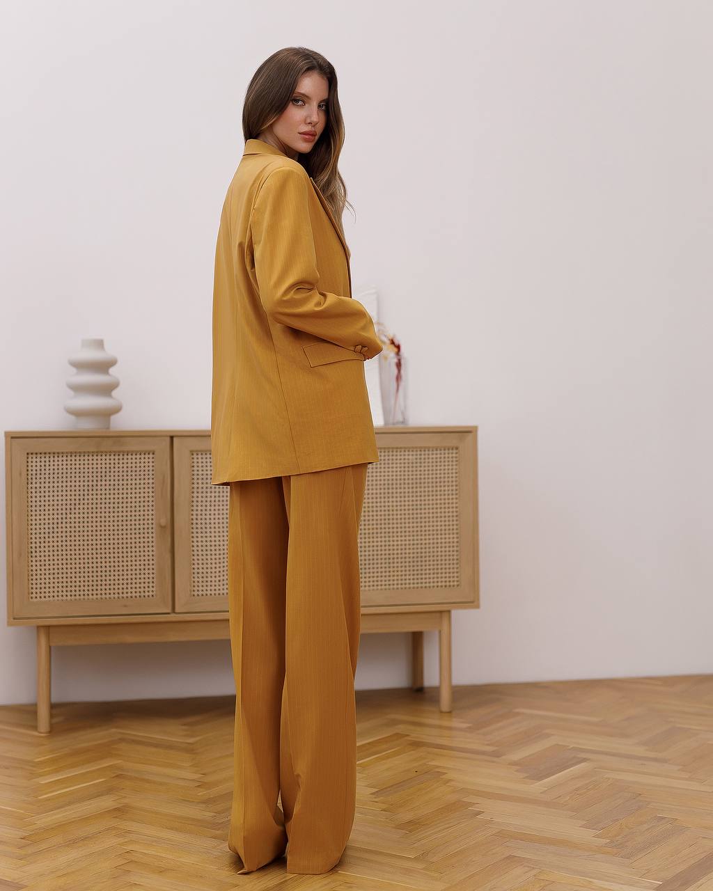 Woman in a mustard yellow outfit standing in a room with wooden flooring and a cabinet.