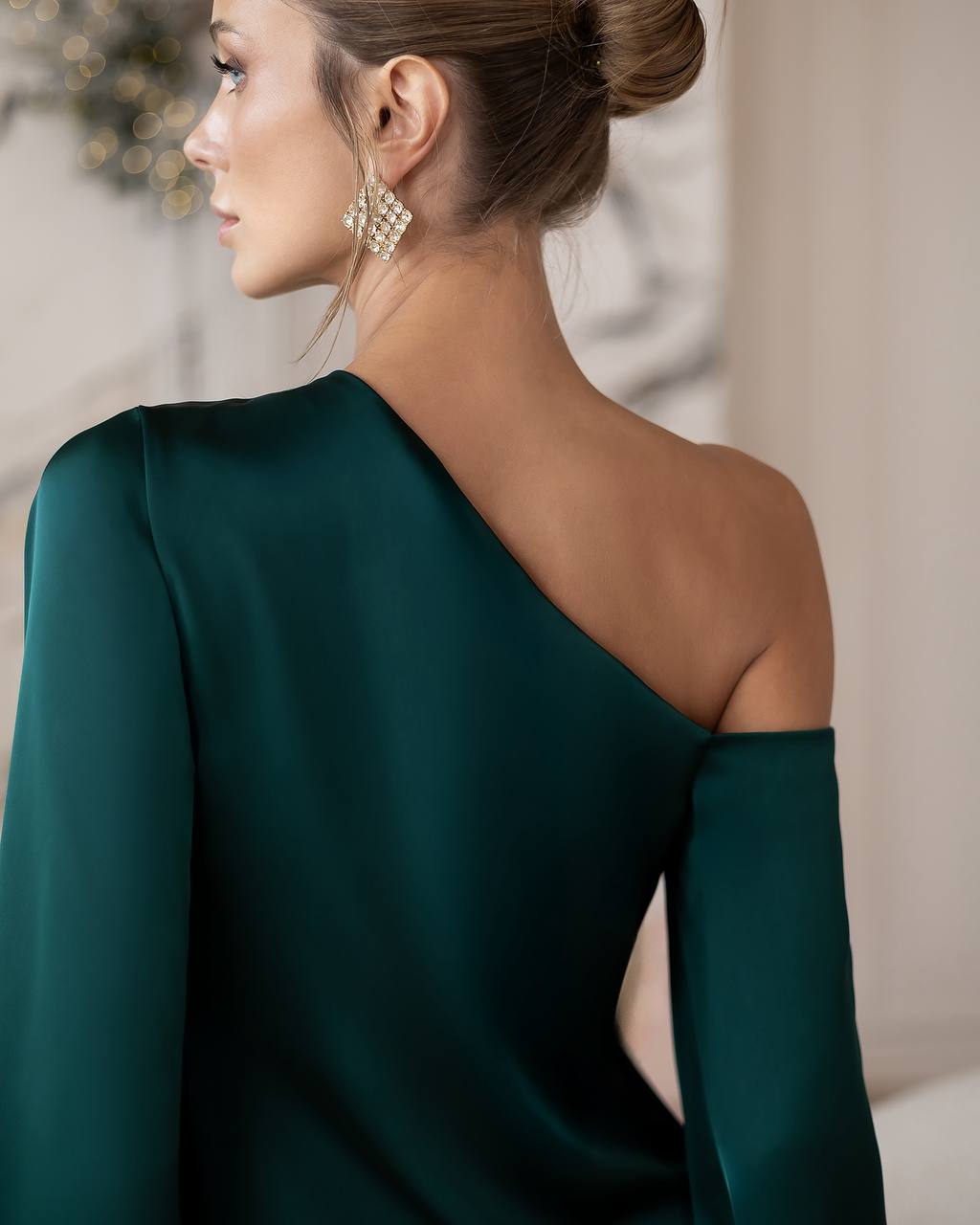A woman shown from the back wearing a dark emerald green one-shoulder dress with a long draped sleeve and a high neckline. She is also wearing a large, textured gold statement earring. The background is a softly blurred indoor setting with warm neutral tones.