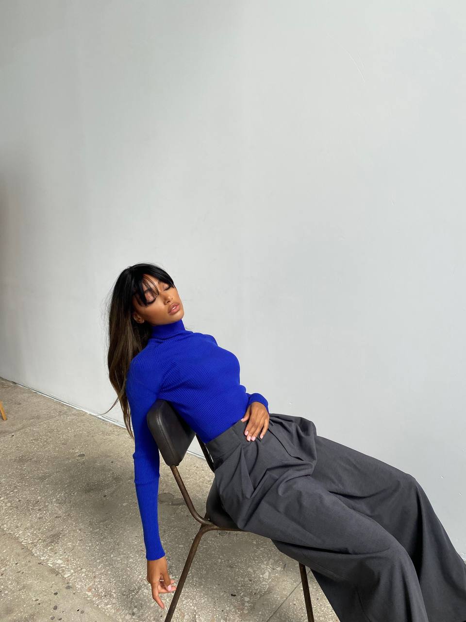 Woman in a blue top and gray pants sitting on a chair against a plain wall.