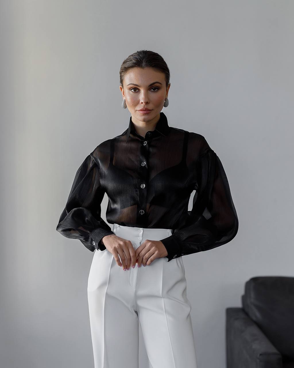 Woman wearing a black sheer blouse and white pants against a gray background