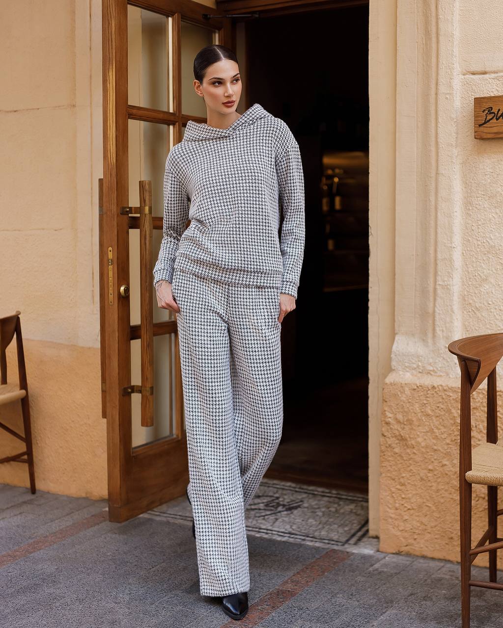 Woman in a checkered outfit standing in a doorway with chairs and a door in the background.