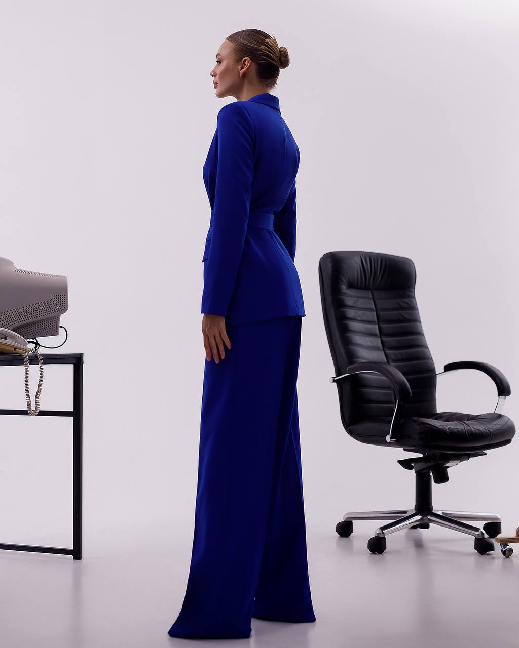Woman in a blue suit standing next to a black office chair on a white background