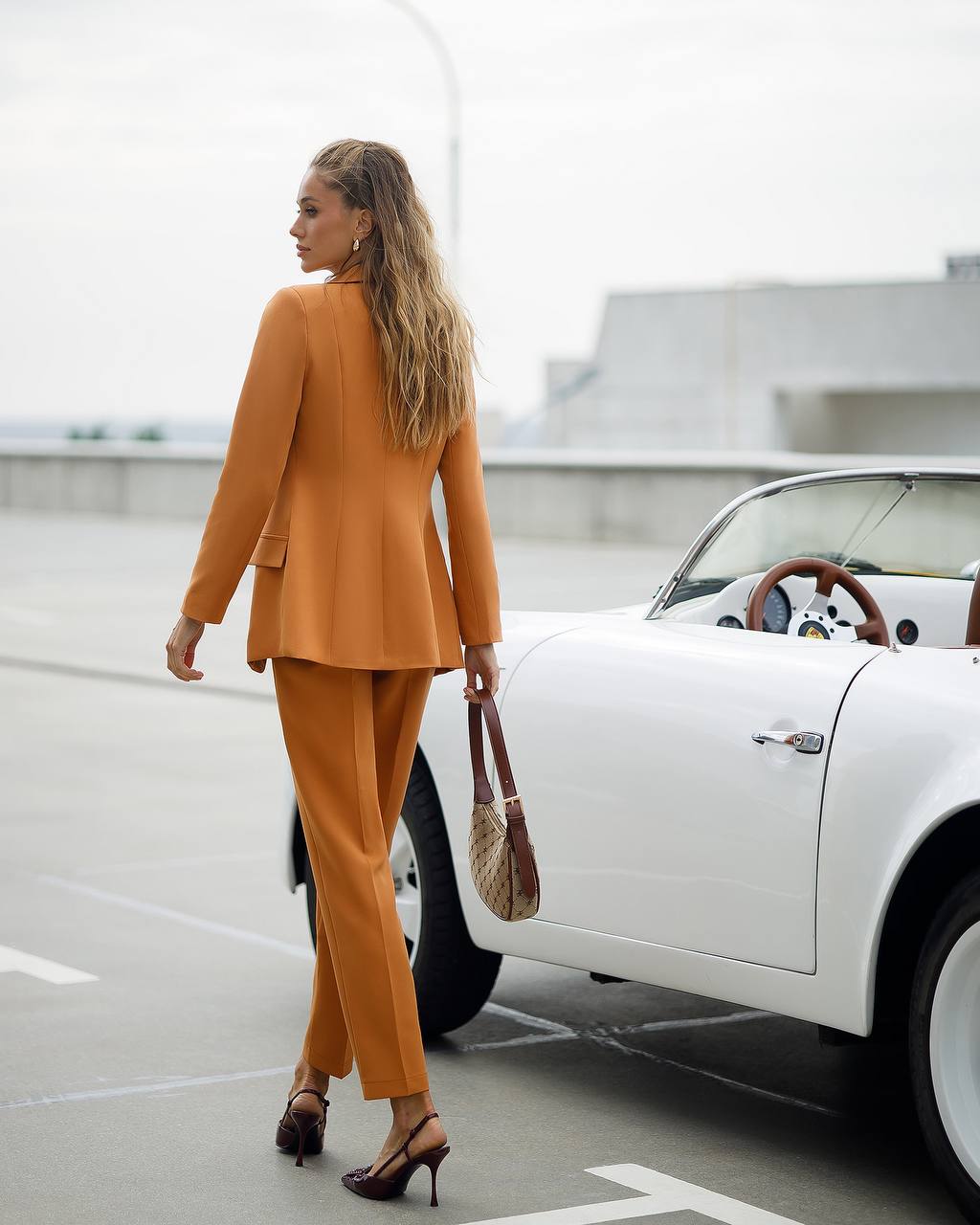 Woman in a mustard suit standing next to a white car on a rooftop.